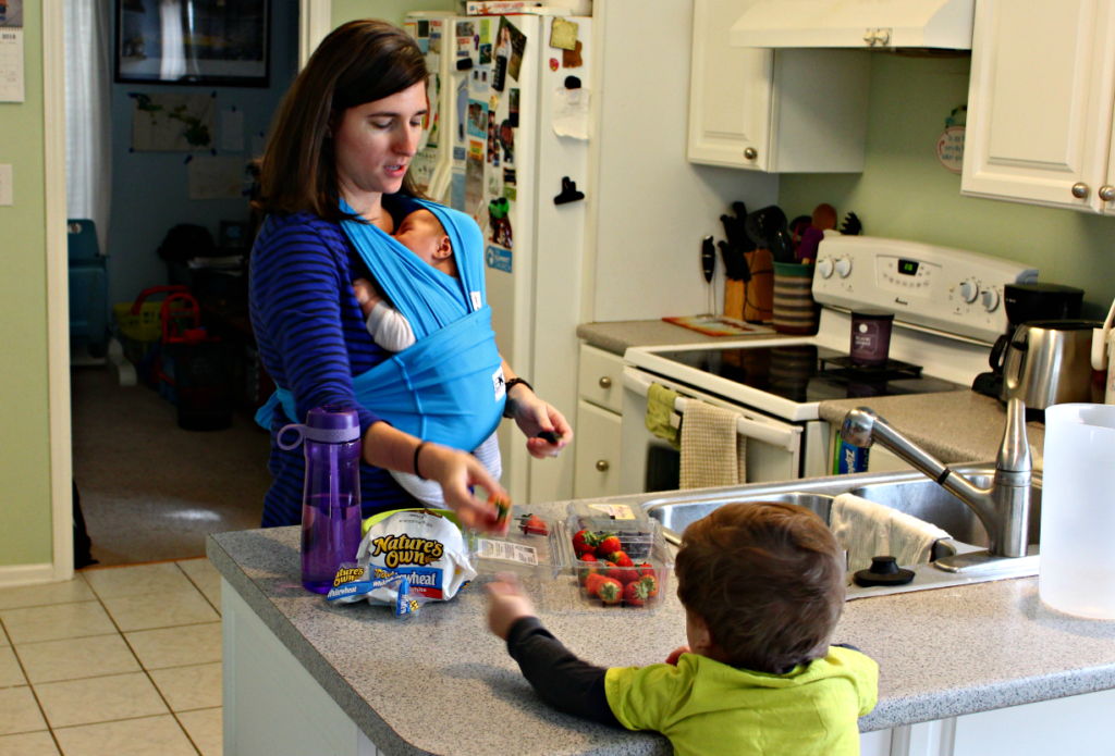 Mother preparing lunch for toddler with infant in a baby carrier with Baby K'tan Active Baby Carrier | from Live Well Play Together Blog | #momlife #motherhood #parenting #babywearing
